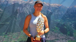 Eine Frau posiert mit einem goldenen Pokal vor einer Kulisse von verschneiten Bergen und einer Stadt unten.
