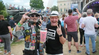 Zwei Männer bei einer Outdoor-Veranstaltung. Einer mit Hut, Bandana und vielen Abzeichen. Ein anderer mit einem rosa Becher und einem schwarzen T-Shirt. Ein großes Gebäude im Hintergrund.