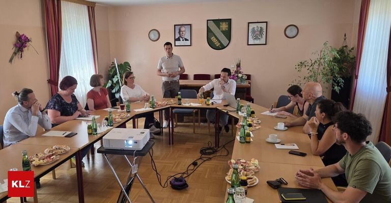 Eine Gruppe von Menschen sitzt um einen Tisch mit Essen und Getränken. Ein Beamer ist auf einem Ständer aufgestellt. Ein Mann steht vor ihnen. Bilder hängen an der Wand.
