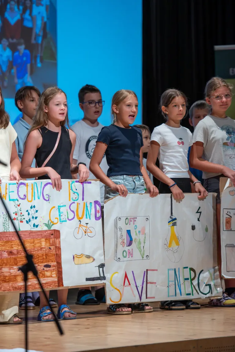 A group of children holding handmade signs with messages like 'Movement is Health' and 'Save Energy' are standing in a row on stage.