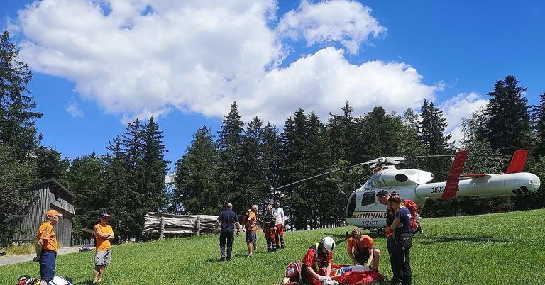 A group of people on a grassy hill assist a person in red who is lying on a red blanket, with a helicopter nearby.