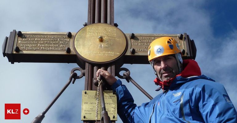 A man in a yellow helmet stands next to a metal cross with plaques, one reading '200 Jahre'. The sky is cloudy.