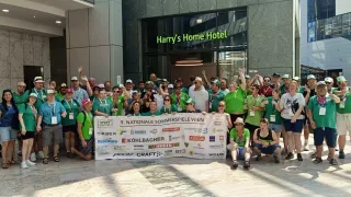 Eine Gruppe von Menschen in grünen Shirts steht vor einem Gebäude und hält ein Banner für die 9. Nationale Sommerspiele Wien.