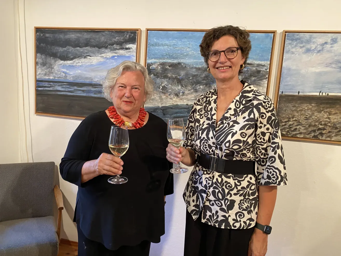 Two women, one wearing a red necklace, stand in front of three paintings, holding wine glasses, smiling.