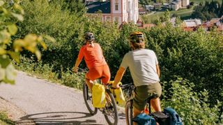 Zwei Radfahrer mit Helmen fahren auf einer kurvenreichen Straße, umgeben von grünen Bäumen und einer Kirche in der Ferne.
