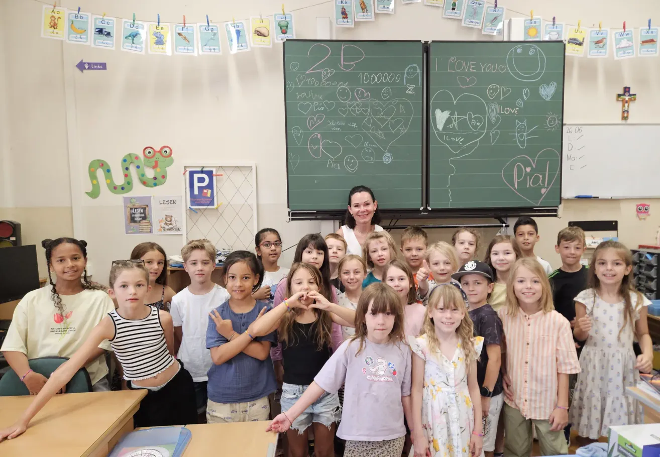 A group of young students and a woman are standing in front of two blackboards. They are all smiling and posing for a picture. The blackboards have drawings of hearts and numbers. The classroom has posters, a whiteboard, and a wooden desk.