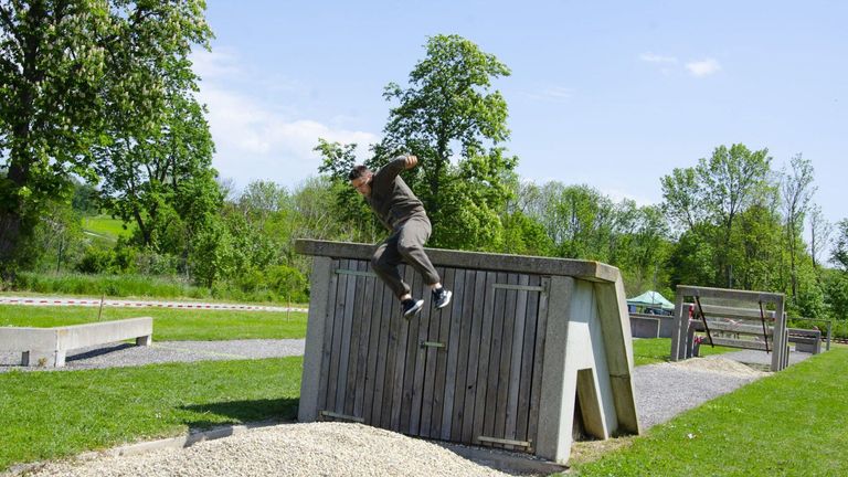 A man in a gray shirt jumps over a wooden structure with a slanted roof, surrounded by trees and a gravel field.