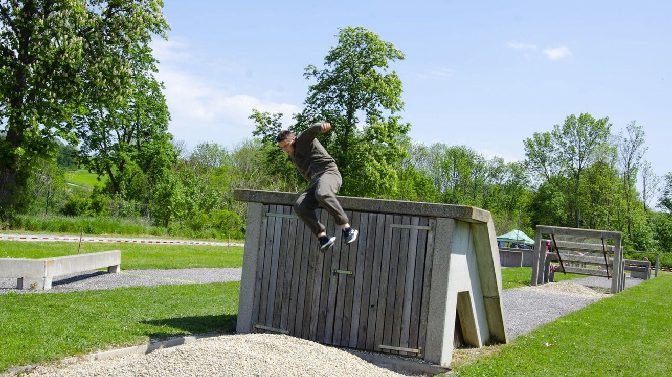 A man in a gray shirt jumps over a wooden structure with a slanted roof, surrounded by trees and a gravel field.
