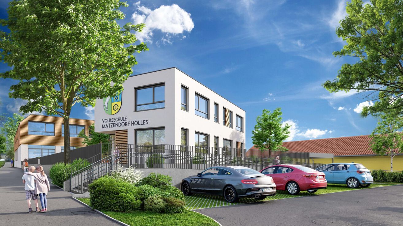 Ein modernes weißes Gebäude, Volksschule Matzendorf Holles, steht mit einem Logo an der Fassade. Autos sind davor geparkt. Bäume und Büsche sind drumherum. Der Himmel ist blau mit Wolken.
