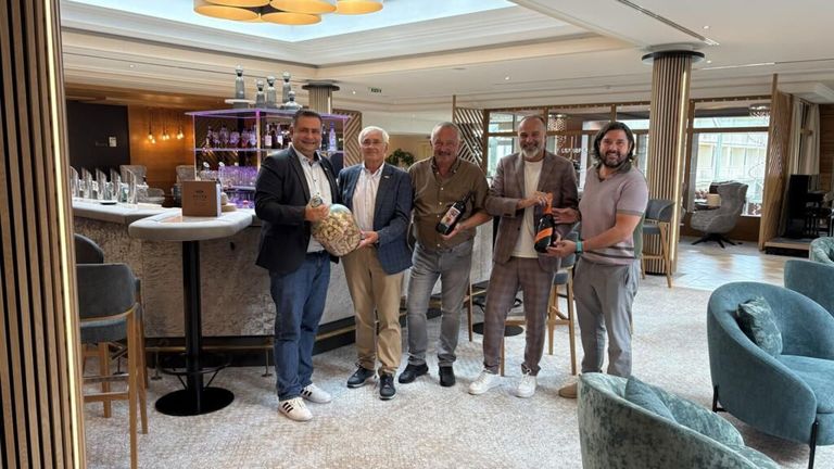 Five men stand indoors, smiling, holding wine bottles and a large jar. Behind them is a bar with shelves of liquor bottles and a counter.