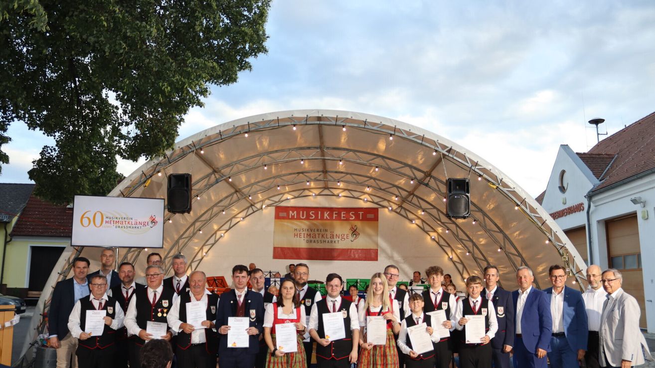 A group of people holding certificates stands in front of a stage, adorned with a banner that reads 'Musikfest Helmatklange Grossmarkt.' They are under a tent with speakers. The sky is cloudy.