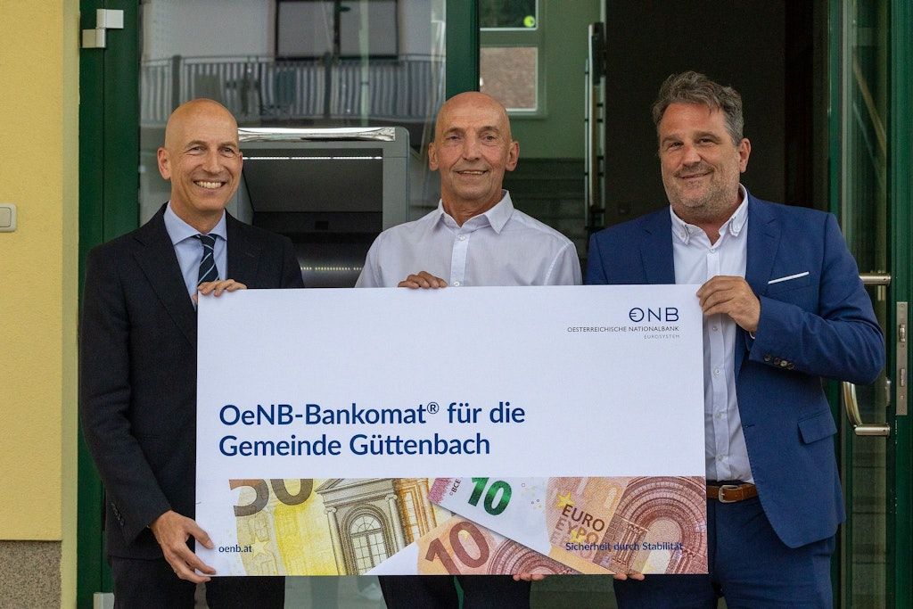 Drei Männer halten ein großes Schild mit einem Banklogo und Bargeld. Sie lächeln und stehen vor einem Geldautomaten. Das Schild trägt die Aufschrift 'OeNB-Bankomat für die Gemeinde Guttenbach'.