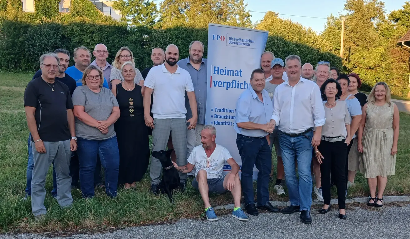 A group of people pose for a picture outside with a sign that says Heimatverpflichtung. A man sits with a dog.