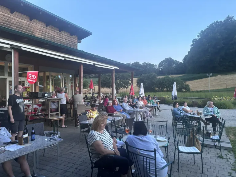 An outdoor restaurant with several people sitting around tables under umbrellas, with a red sign on the building.