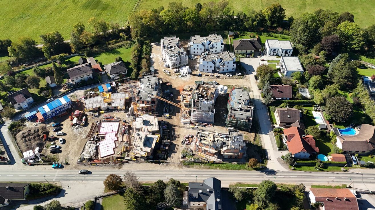An aerial view of an ongoing construction site with several buildings under construction, some completed, and others still in progress. There are trees and grass in the surrounding area.