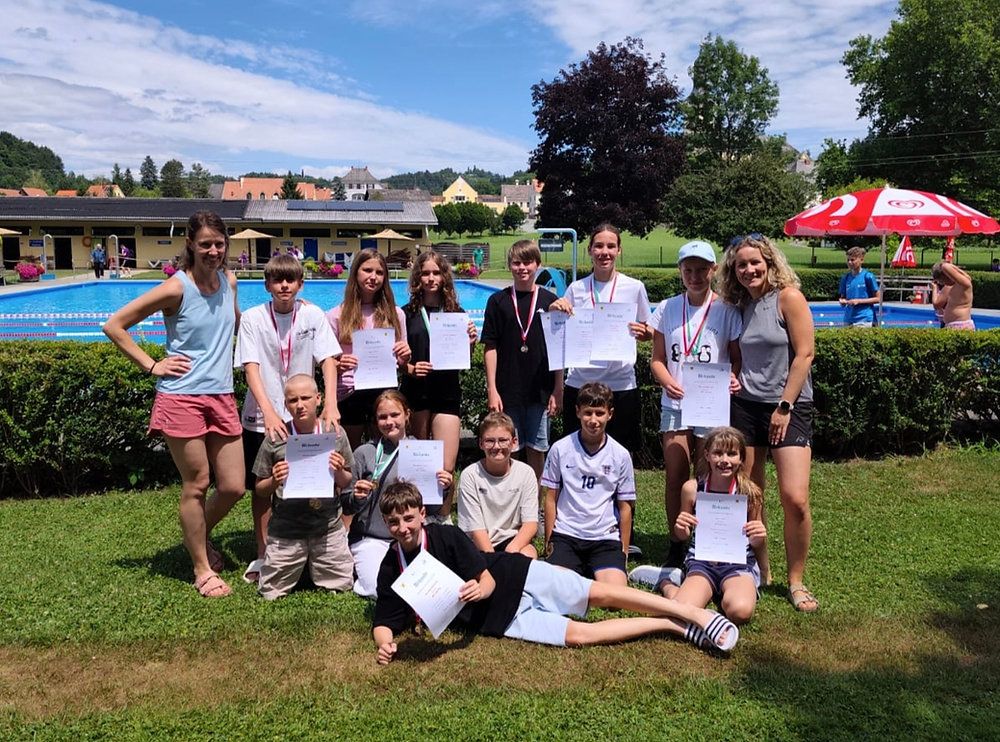 A group of young swimmers with medals and certificates pose for a photo by a pool.