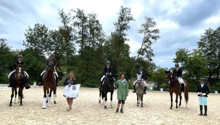 A group of people and horses stand on a sandy field. The riders wear helmets and gloves, and a woman holds a blanket. The horses are saddled, and the area is surrounded by trees.