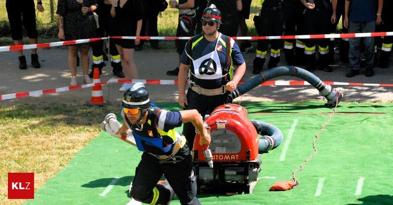 A man wearing a safety suit operates a red machine while another man in a helmet watches on a green field with onlookers behind barriers.
