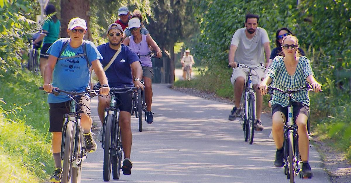 A group of cyclists riding on a paved road, surrounded by greenery. Some wear hats, glasses, and sneakers. Two cyclists in front, one with a backpack.