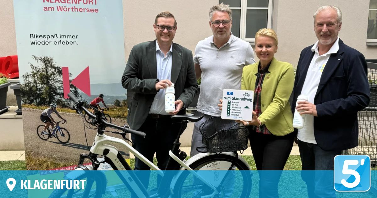 Four individuals stand beside a bicycle with a basket, one holding a sign reading Zum Glanradweg and another holding a bottle. Behind them, a cyclist rides on a path.