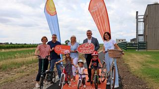 Eine Gruppe von Menschen posiert für ein Foto auf einem Fahrradweg mit Fahnen dahinter. Sie halten Schilder mit den Aufschriften 'Burgenland radelt' und 'Rad frei'.