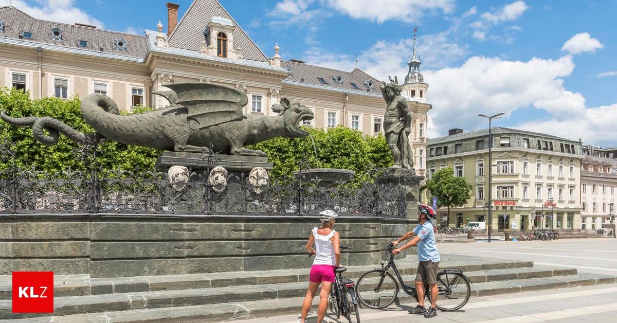 Two cyclists pose for a photo in front of a dragon fountain in a historic town square.