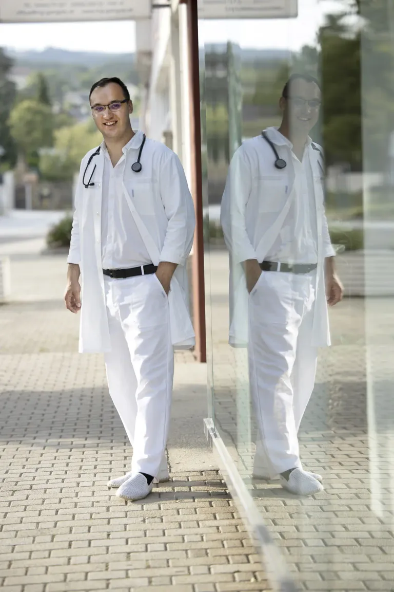 A smiling man dressed in white medical attire walks on a brick path with a stethoscope around his neck, reflected in a glass.