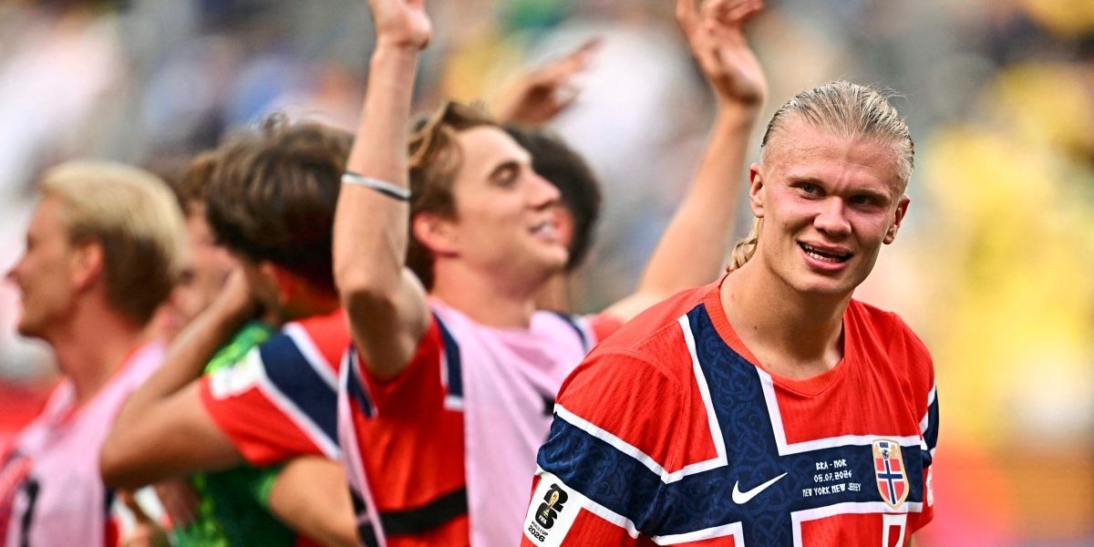 Ein Fußballspieler in einem rot-blauen Trikot mit Nike-Logo steht lächelnd. Andere Spieler heben im Hintergrund die Hände.