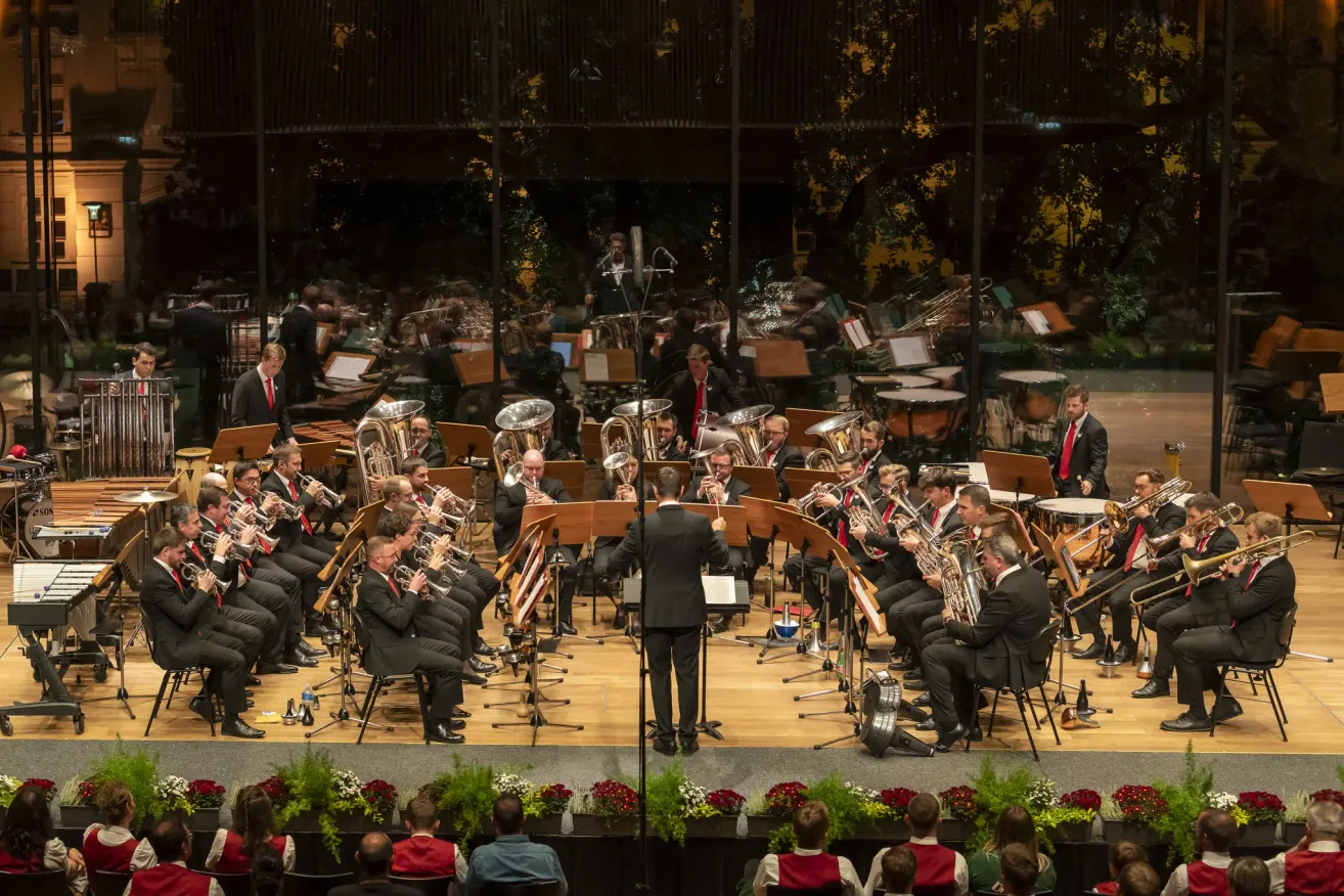 Ein Orchester spielt auf der Bühne. Die Musiker sitzen und stehen und spielen verschiedene Instrumente. Ein Dirigent steht vor ihnen. Das Publikum sitzt vorne und schaut zu.