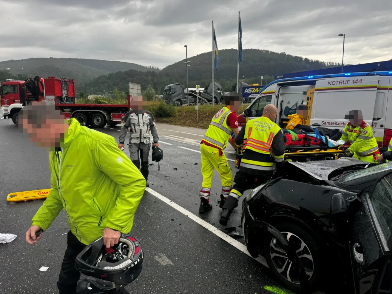 Emergency responders assist individuals near a wrecked vehicle on the roadside, with a damaged truck and ambulance in the background.