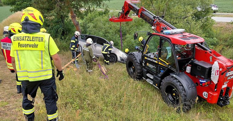 Feuerwehrleute arbeiten mit einem Kran und einem Traktor an der Entfernung eines Autos aus einem Baum, wobei eine Person ein Werkzeug in einem Grasbereich hält.
