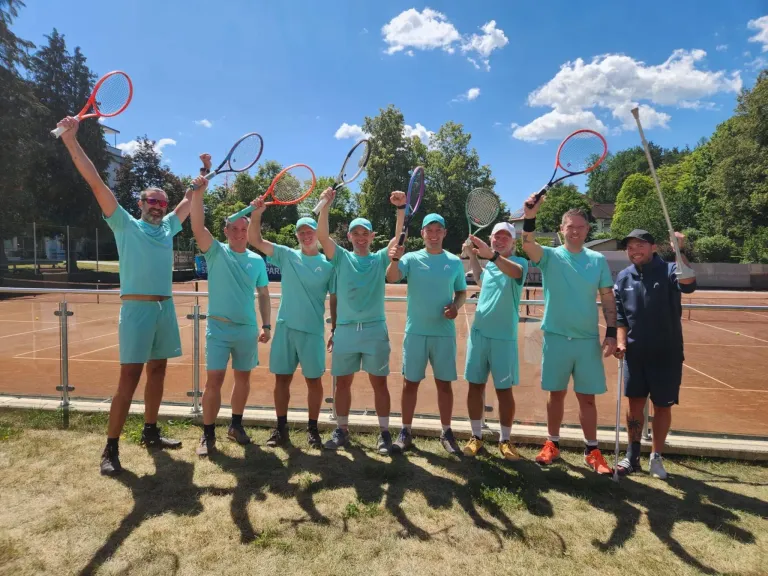 A group of men in matching turquoise outfits are posing for a photo with tennis rackets on a grassy field. They are standing on a tennis court, with a fence and trees in the background.