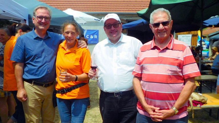 Vier Menschen stehen bei einem Event und lächeln. Eine Frau hält ein Weinglas, und ein Mann hält ein Glas Wein. Sie stehen unter einem Zelt.