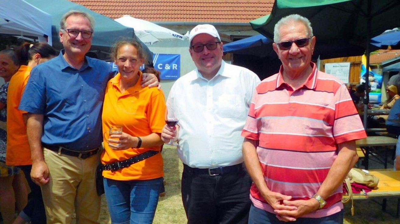 Vier Menschen stehen bei einem Event und lächeln. Eine Frau hält ein Weinglas, und ein Mann hält ein Glas Wein. Sie stehen unter einem Zelt.