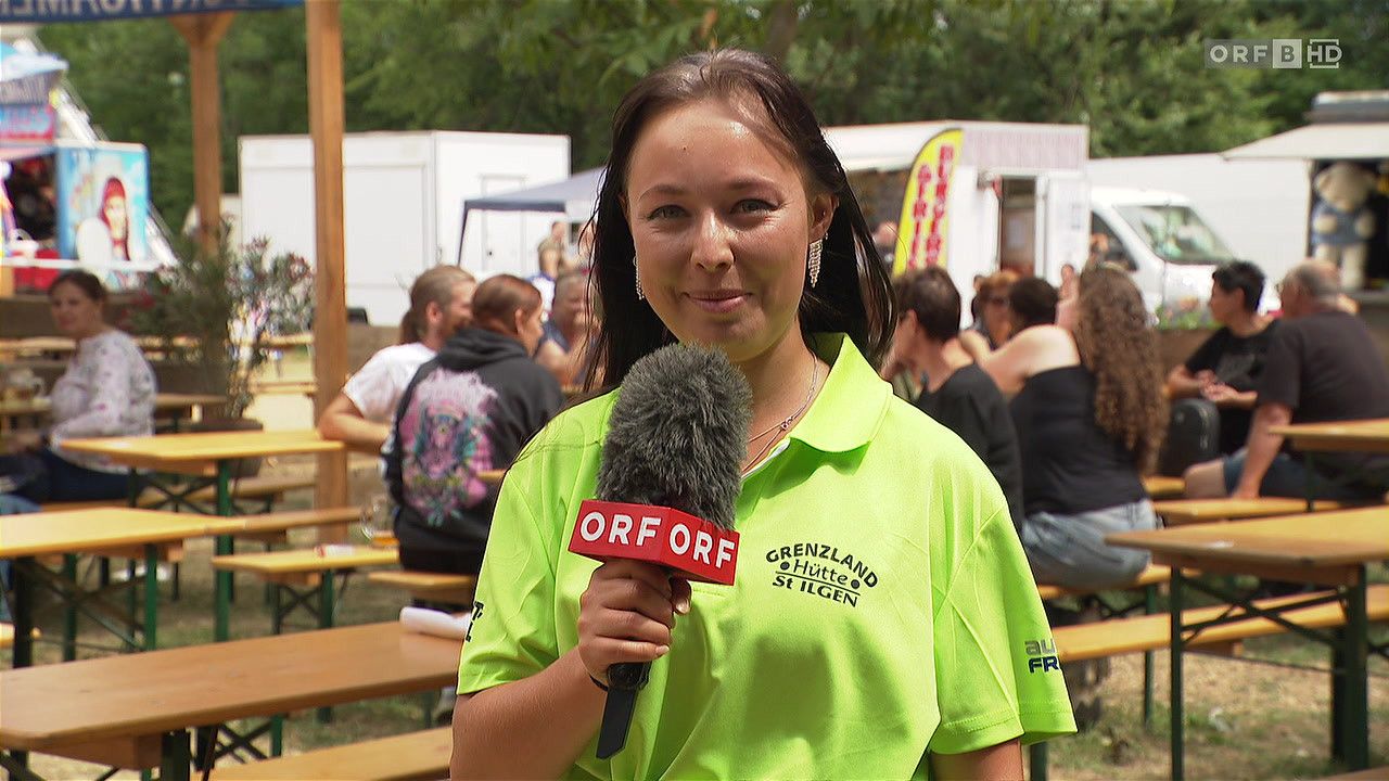 A woman wearing a neon green shirt with the words ORF on it is holding a microphone and smiling. Behind her are people sitting on benches.