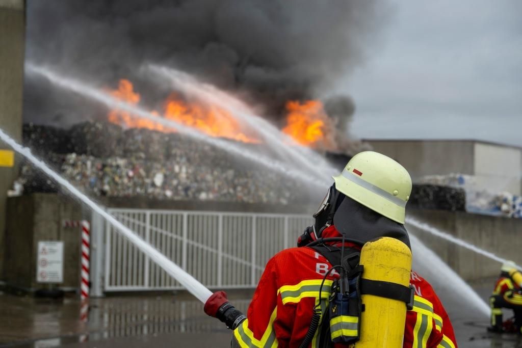 Ein Feuerwehrmann in voller Ausrüstung, einschließlich Helm und Maske, spritzt Wasser aus einem Schlauch auf ein großes Feuer, das in einem Haufen Schutt brennt.
