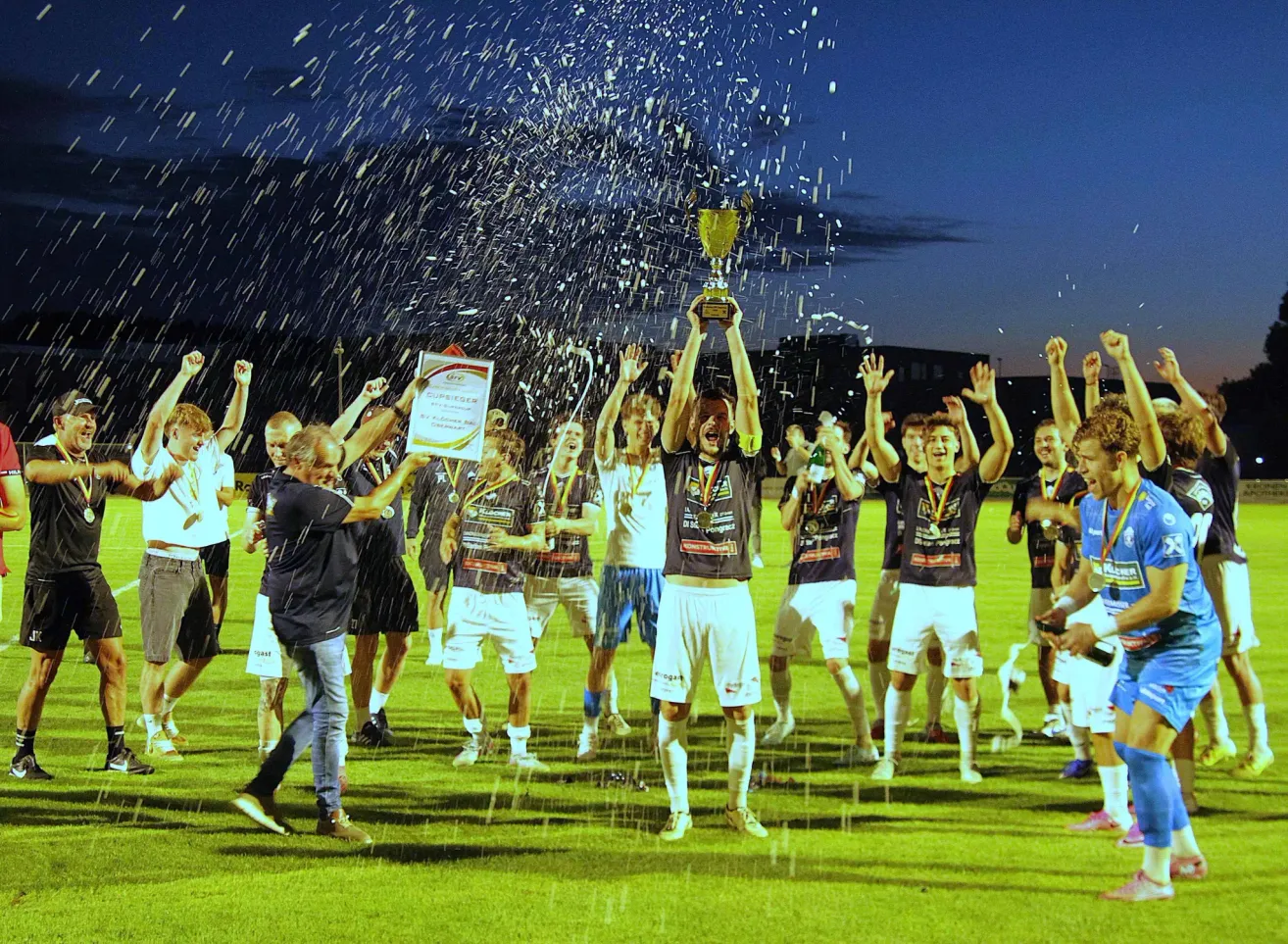 Eine Fußballmannschaft feiert ihren Sieg auf einem Feld mit Konfetti und einem Pokal. Spieler und Trainer heben die Hände in Freude.