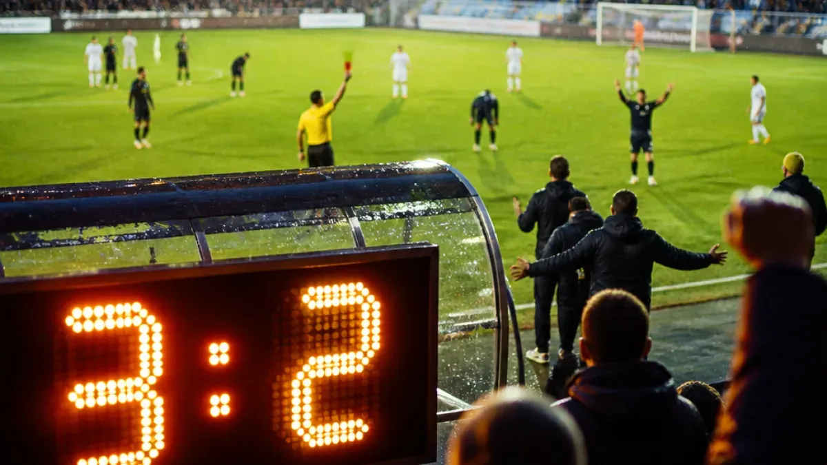 Fußballspieler auf einem Feld, ein Schiedsrichter zeigt eine rote Karte und eine Anzeigetafel zeigt 1:2.