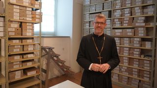 Ein Priester steht in einem Raum mit Regalen voller Schachteln hinter ihm, trägt einen schwarzen Umhang und eine goldene Kreuzkette. Er lächelt und scheint für ein Foto zu posieren.