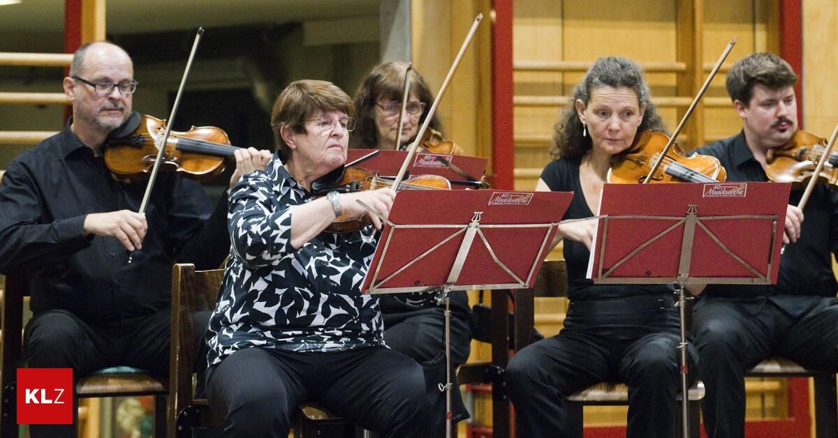 Eine Gruppe von Frauen spielt Violinen in einem Konzertsaal. Sie sitzen auf Stühlen mit Violinständern vor ihnen. Sie tragen alle Brillen und scheinen sich auf ihre Aufführung zu konzentrieren. Die Frau in der Mitte hält einen Geigenbogen.