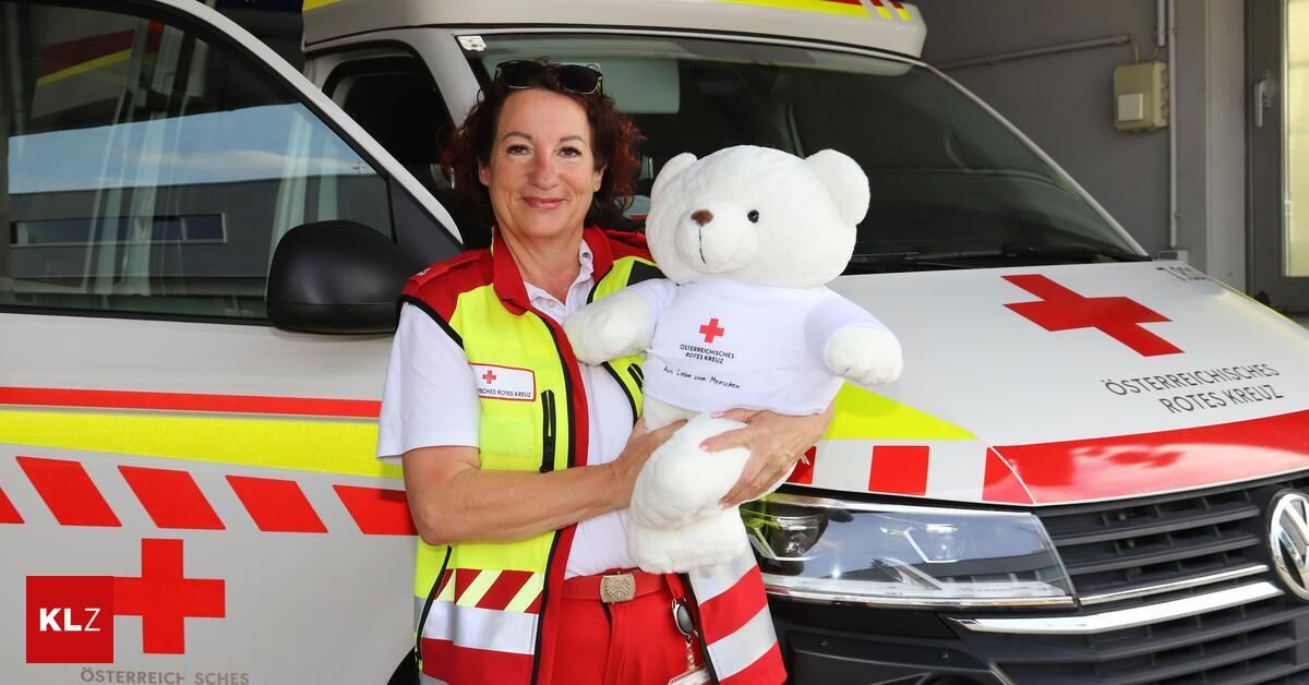 A smiling woman in a red and white uniform holds a white teddy bear with a red cross on its chest in front of an ambulance.