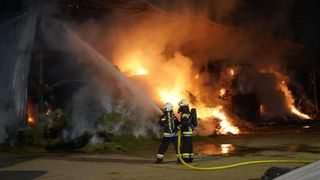 Two firefighters in full gear are battling a large fire at night, spraying water from a hose.