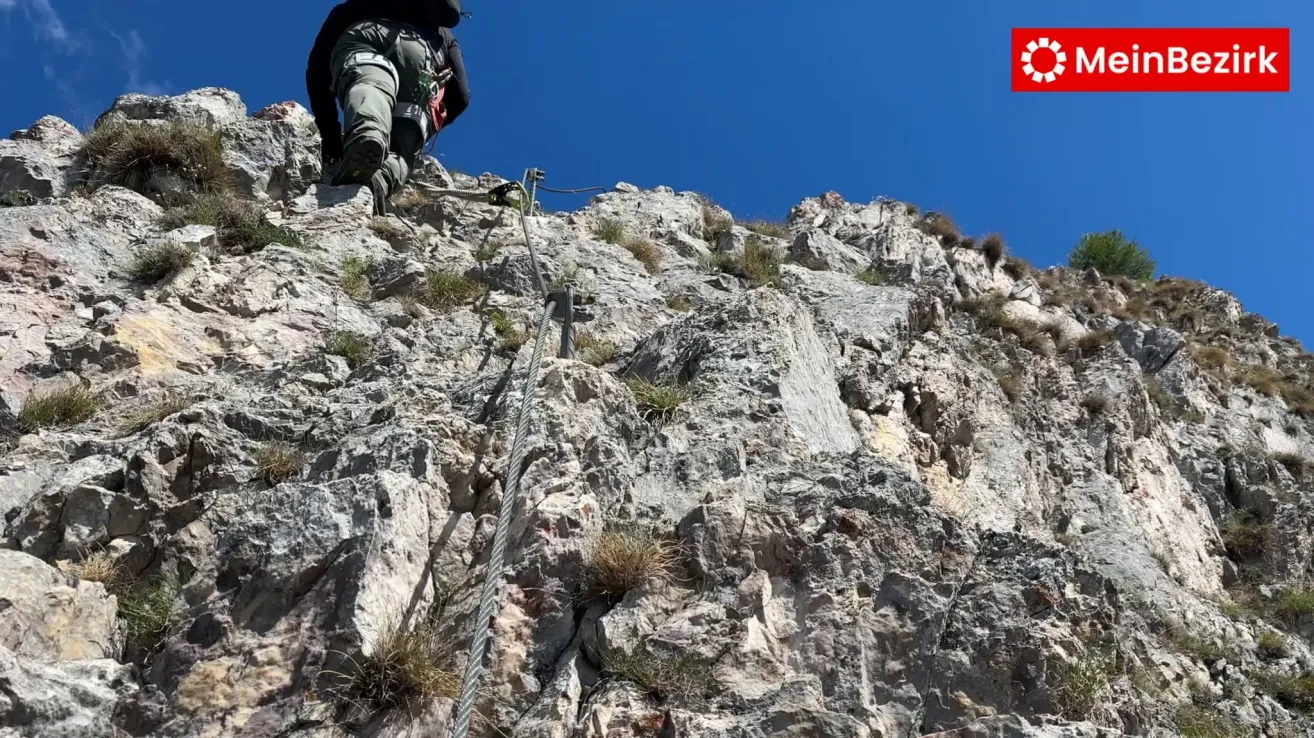 A person is climbing a steep rocky cliff using a rope and harness. The climber is wearing a helmet and harness. The sky is clear and blue.