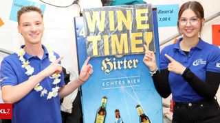 Two people stand next to a blue poster advertising 'Sürter Echtes Bier' with the slogan '4 WINE TIME'. The man wears a garland of white flowers around his neck.