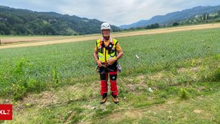 A man in safety gear stands in a grassy field with mountains in the background. He wears a white helmet, a yellow safety jacket, and red and black pants. He is holding a rope and appears to be preparing for an activity.