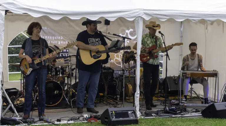 Two men are performing on stage under a tent, one playing a guitar and the other playing a bass guitar. A drum set is behind them.