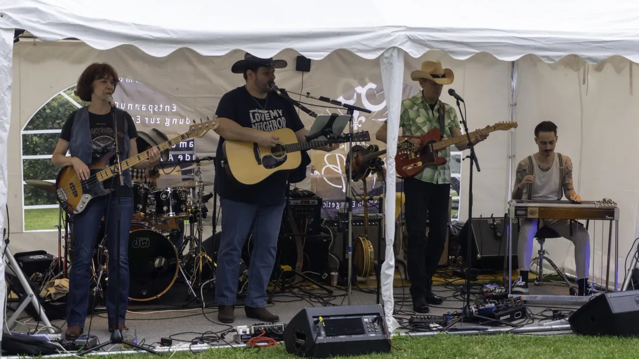 Two men are performing on stage under a tent, one playing a guitar and the other playing a bass guitar. A drum set is behind them.