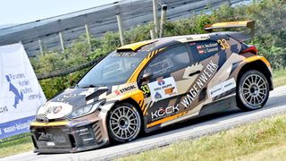 Ein Rallyeauto mit Tarndesign, Nummer 33, fährt auf einer Straße. Es hat Sponsorenlogos an der Seite und vorne.