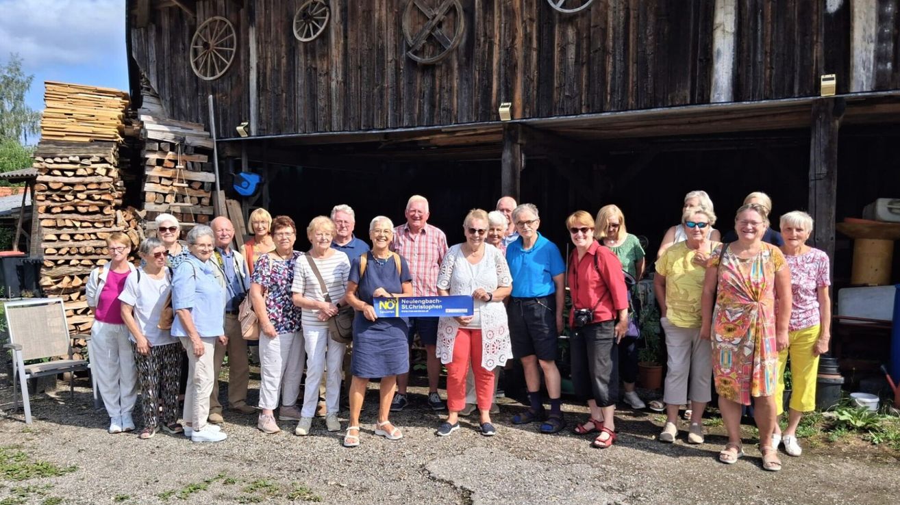 Eine Gruppe älterer Menschen steht vor einem Scheunengebäude, lächelt und posiert für ein Foto. Sie halten ein blaues Banner mit Text.