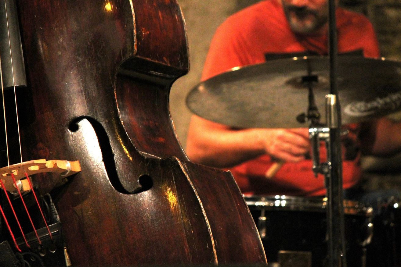 A close-up of a double bass with a musician playing the drums in the background.
